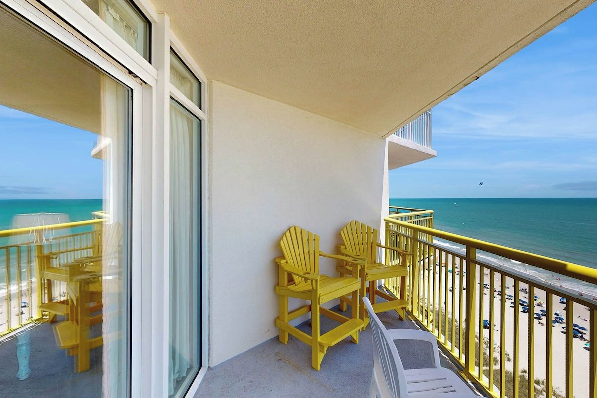 Un balcon avec des fauteuils Adirondack jaunes, une petite table et une vue sur la plage et l’océan.
