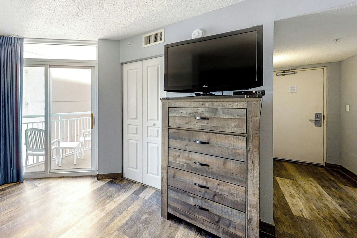 A room with a flat-screen TV on a wooden dresser, a sliding glass door leading to a balcony, and a white door.