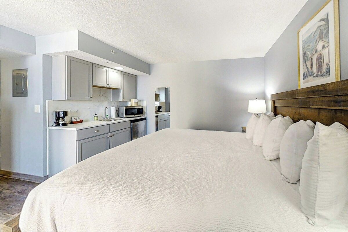 Habitación de hotel con una cama grande, cabecera de madera, una pequeña kitchenette y un cuadro en la pared.