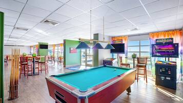 A pool table in a brightly lit room with a green pool table, a pool cue rack, and a billiards table.