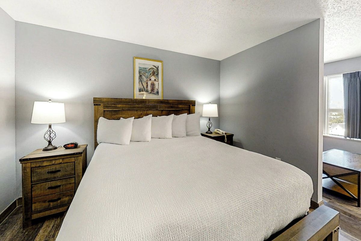 Une chambre d’hôtel avec un grand lit, deux tables de chevet, une petite table, un tableau encadré au mur et une fenêtre avec des rideaux.