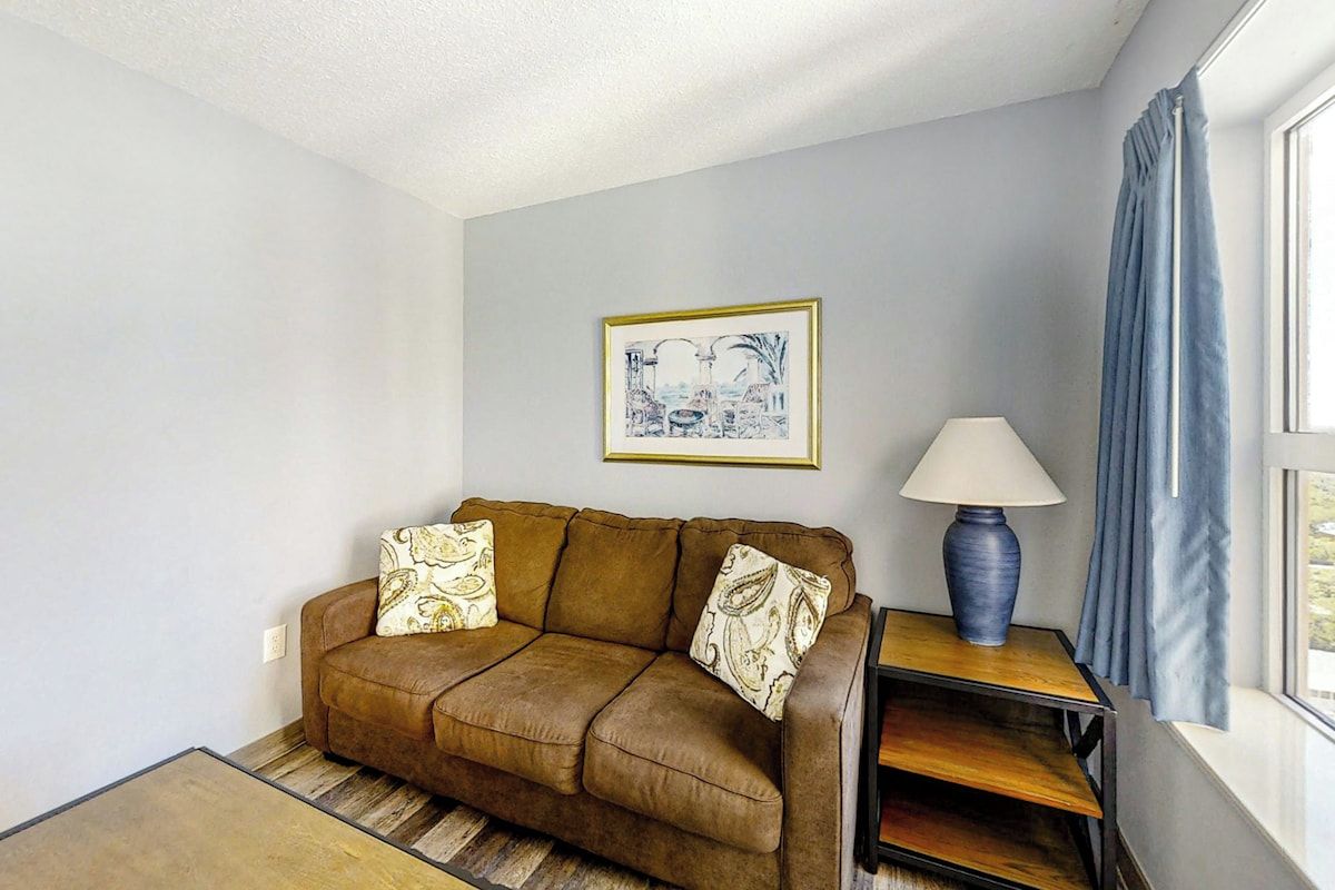 A living room with a brown sofa, patterned cushions, a framed picture on the wall, a lamp on a side table, and a window with curtains.