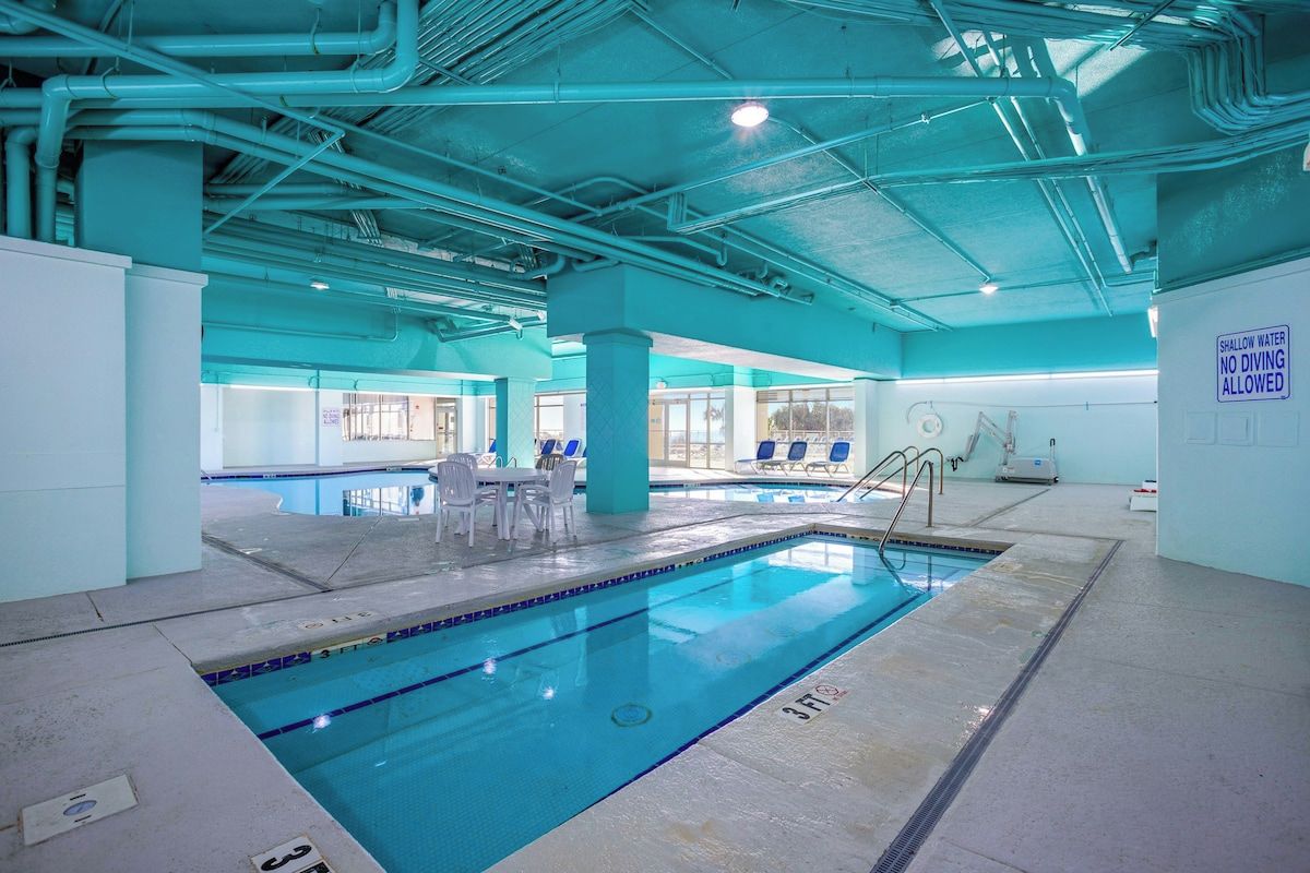 An indoor swimming pool with a shallow water sign, lounge chairs, and a tiled floor.