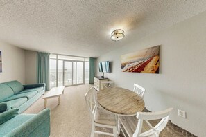 A living room with a sofa, a dining table with chairs, a TV, and a painting on the wall. - Bay Watch Resort 1219 (North Myrtle Beach)