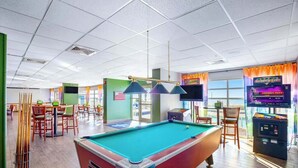 Une table de billard se trouve dans une pièce bien éclairée, avec un porte-queue vert, une autre table de billard et un écran de télévision diffusant un match.