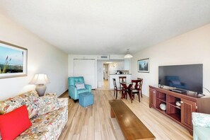 A living room with a sofa, a teal armchair, a wooden bench, a dining table with chairs, a TV on a wooden stand, and a kitchen area in the background. - Bay Watch Resort 1220 (North Myrtle Beach)