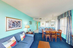 Ein Zimmer mit blauem Sofa, einem Esstisch mit Stühlen, einer Küchenzeile und einem Fenster mit Vorhängen.