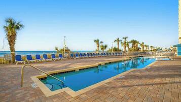 A pool area with lounge chairs and palm trees, a clear sky, and a building in the background.