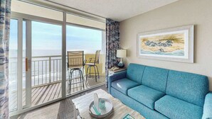 A room with a blue sofa, a table with a lamp, a painting, and a view through a sliding door to the outside. - Bay Watch Resort 1407 (North Myrtle Beach)