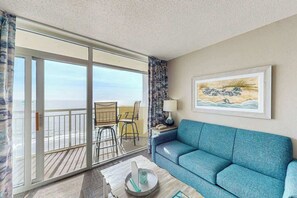 A room with a blue sofa, a table with a lamp, a painting, and a view through a sliding door to the outside. - Bay Watch Resort 1407 (North Myrtle Beach)