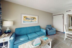 A room with a blue sofa, a patterned armchair, a small table with a lamp, and a framed picture on the wall. - Bay Watch Resort 1407 (North Myrtle Beach)