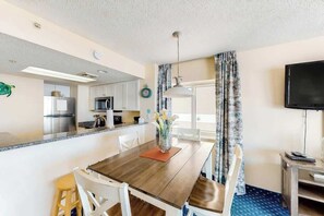 A modern kitchen with stainless steel appliances, a dining area with a wooden table and chairs, and a flat-screen TV mounted on the wall. - Bay Watch Resort 1411 (North Myrtle Beach)