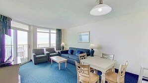 A hotel room with a sofa, a dining table with chairs, a TV, and a balcony with curtains. - Bay Watch Resort 1412 (North Myrtle Beach)