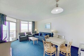 Ein Hotelzimmer mit einer Sitzecke, einem Esstisch mit Stühlen, einem Fernseher und einem Balkon mit Vorhängen.