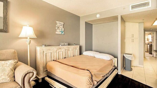 A bedroom with a bed, bedside table, lamp, and a dresser. - Bay Watch Resort 1414 (North Myrtle Beach)
