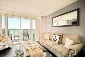 A living room with a sofa, a coffee table, and a balcony with chairs. - Bay Watch Resort 1414 (North Myrtle Beach)