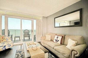A living room with a sofa, a coffee table, and a balcony with chairs. - Bay Watch Resort 1414 (North Myrtle Beach)