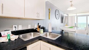 A modern kitchen with two sinks, a black countertop, and white cabinets. There is a window with a view of the ocean. - Bay Watch Resort 1414 (North Myrtle Beach)