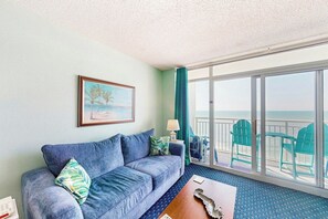 A room with a blue sofa, a painting, a lamp, and a view of the ocean through large windows. - Bay Watch Resort 1419 (North Myrtle Beach)