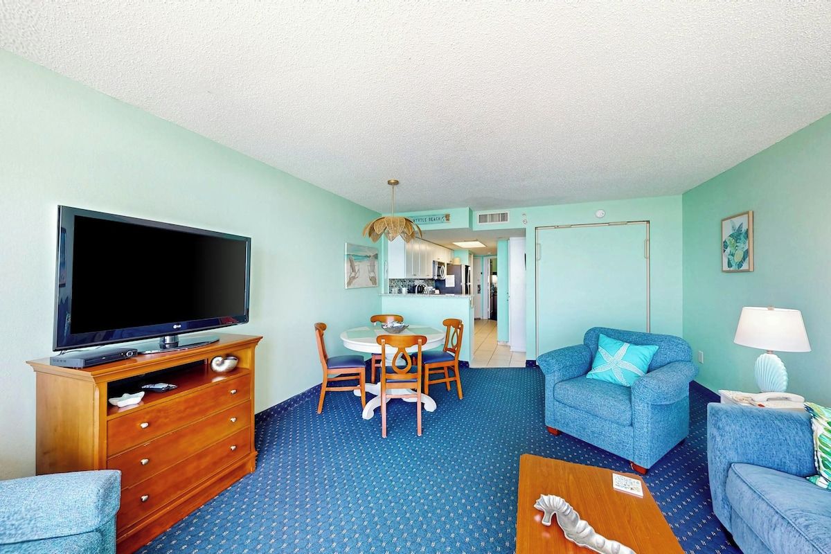 A room with a television, wooden dresser, blue patterned carpet, a dining table with chairs, a blue sofa, and a lamp.