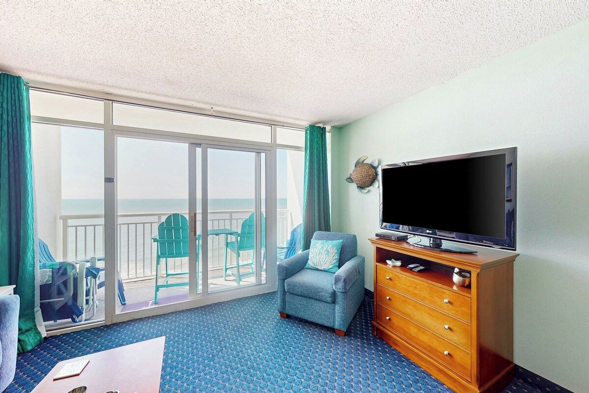 Quarto de hotel com uma grande porta de vidro deslizante que dá para uma varanda, uma televisão sobre um suporte de madeira e um tapete azul com padrão.
