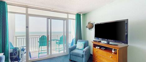 Quarto de hotel com uma grande porta de vidro deslizante que dá para uma varanda, uma televisão sobre um suporte de madeira e um tapete azul com padrão.