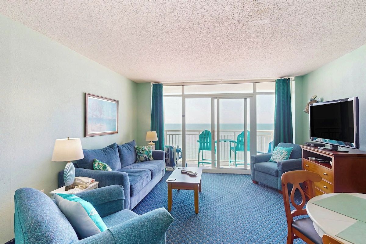 A hotel room with a sofa, a television, a table, and a balcony with chairs.