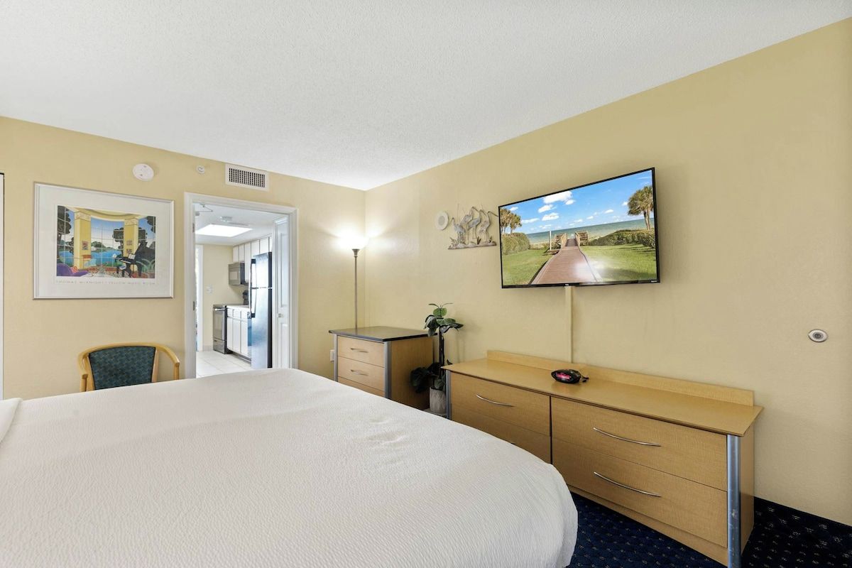 A hotel room with a large bed, a desk with drawers, a television mounted on the wall, and a door leading to a bathroom.