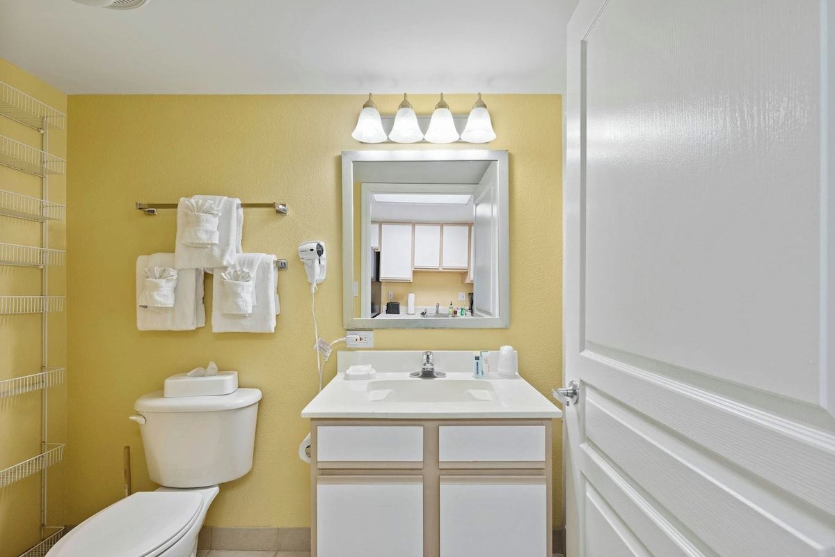A bathroom with a toilet, sink, and mirror. There are towels hanging on a rack, and a shower area is visible.