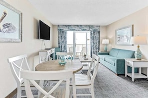 A hotel room with a dining area, a sofa, a TV, and a balcony with curtains. - Bay Watch Resort 1539 (North Myrtle Beach)