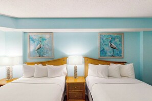 A hotel room with two beds, each with white bedding and a wooden headboard. There are two wall-mounted paintings of a bird, and a nightstand with a lamp between the beds. - Bay Watch Resort 1605 (North Myrtle Beach)