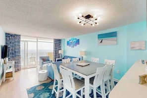A modern living room with a dining area, a large window, and a balcony. - Bay Watch Resort 1605 (North Myrtle Beach)