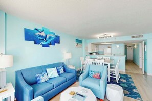 A living room with a blue sofa, a coffee table, and a dining area with white chairs and a table. - Bay Watch Resort 1605 (North Myrtle Beach)