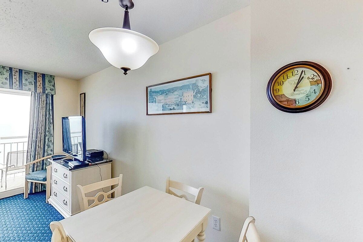 A hotel room with a dining table, chairs, a television on a dresser, a clock on the wall, and a window with curtains.