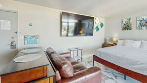 A hotel room with a bed, a brown leather sofa, a small side table, a wall clock, and artwork. - Bay Watch Resort 1501 (North Myrtle Beach)