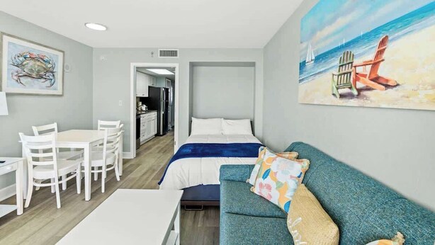 A room with a bed, a blue sofa, a dining table with chairs, and a painting of a beach scene. - Bay Watch Resort 1506 (North Myrtle Beach)