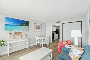 A modern living room with a dining area, a kitchen in the background, and a large beach-themed painting on the wall. - Bay Watch Resort 1506 (North Myrtle Beach)