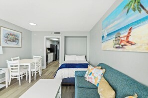 A hotel room with a bed, a sofa, a dining table with chairs, and a painting of a beach scene. - Bay Watch Resort 1506 (North Myrtle Beach)