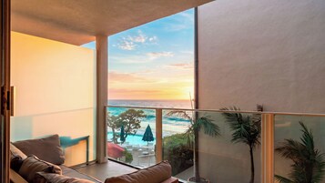 A balcony with a view of the ocean at sunset, a pool area with umbrellas, and a seating area with cushions.