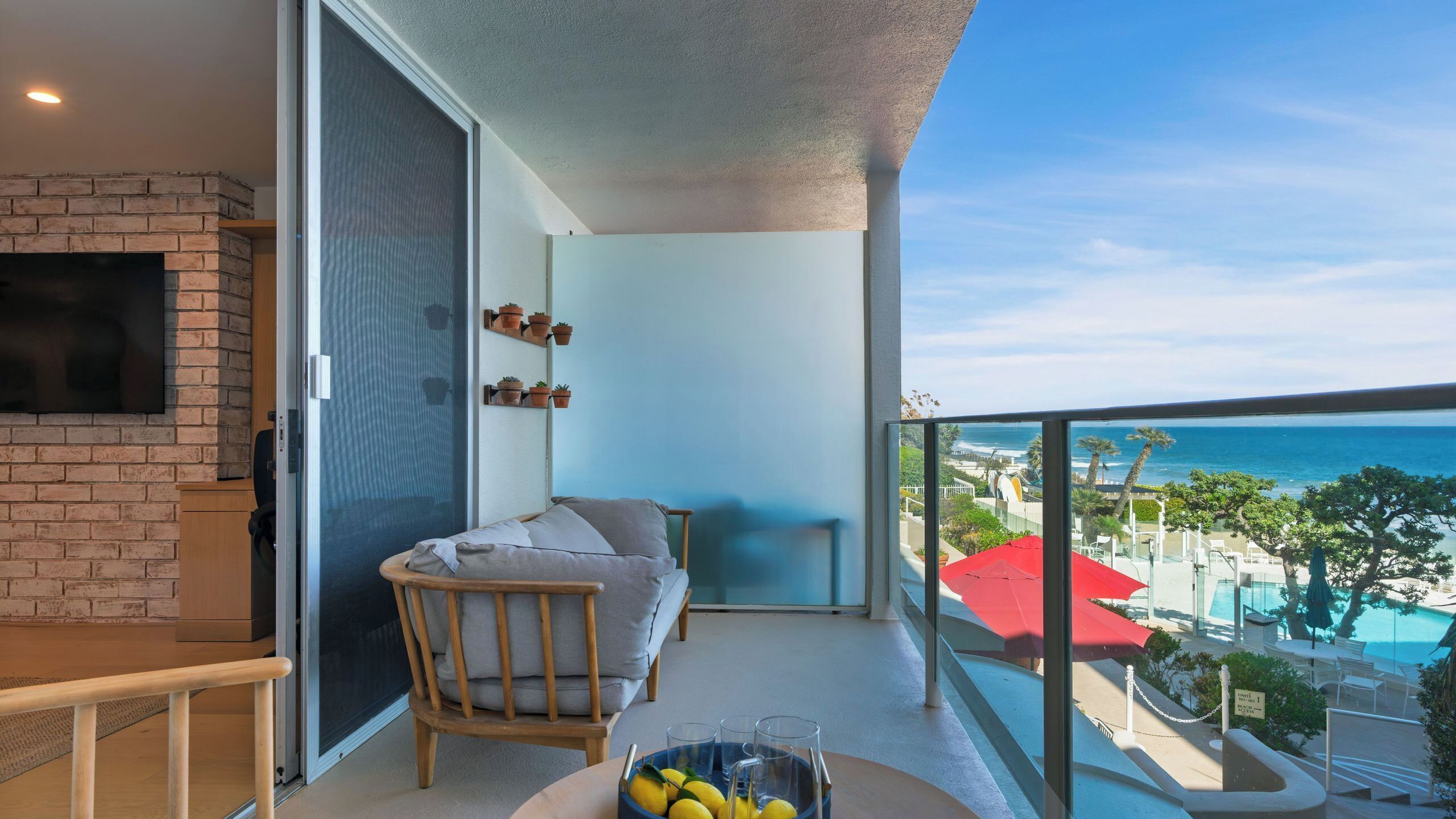 A balcony with a view of the sea, a sofa, a TV mounted on a brick wall, and a table with lemons and glasses.
