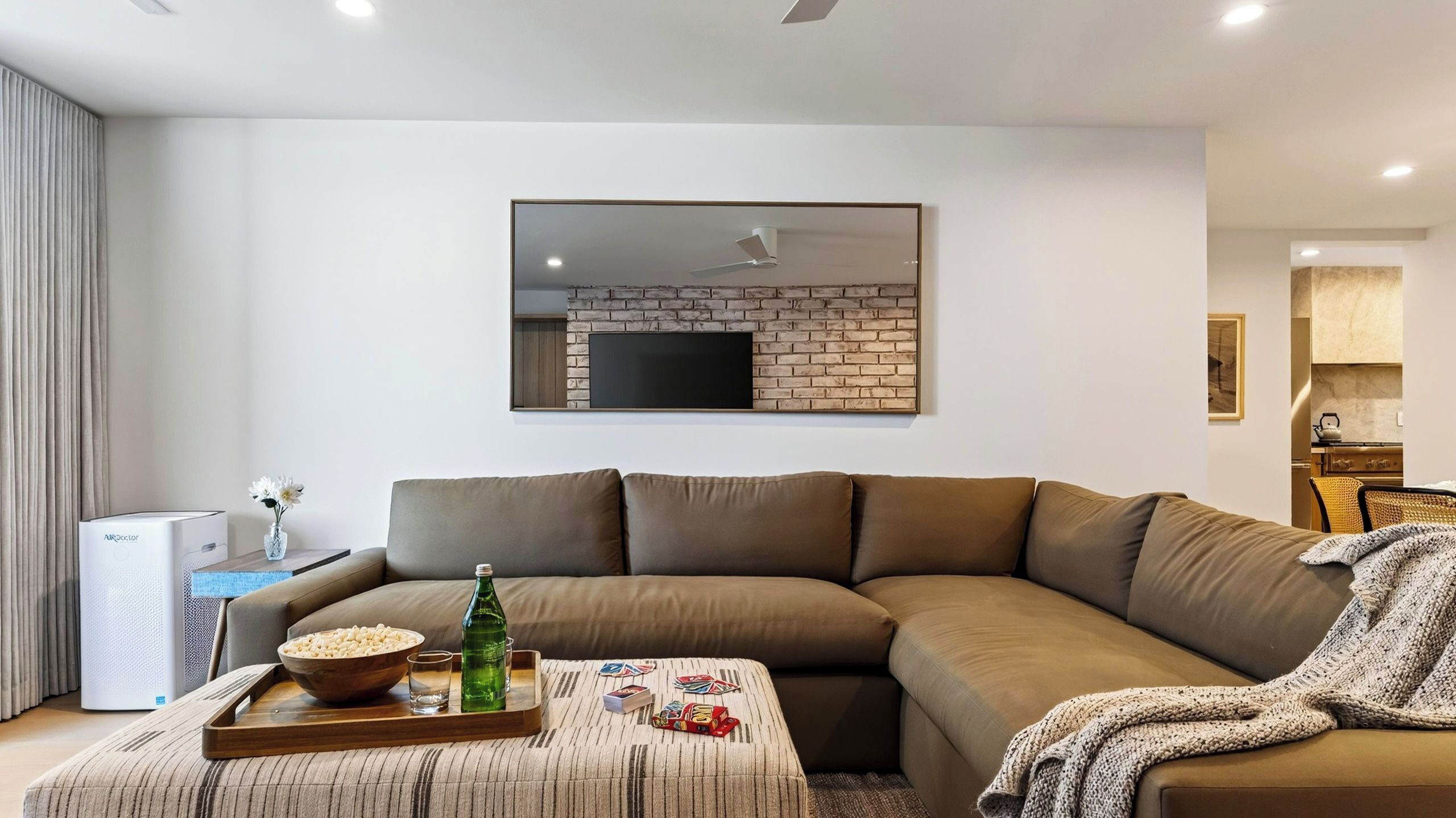 Un salon moderne avec un canapé d’angle, un téléviseur à écran plat fixé à un mur en briques, et une petite cuisine en arrière-plan.