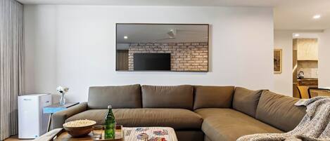 Un salon moderne avec un canapé d’angle, un téléviseur à écran plat fixé à un mur en briques, et une petite cuisine en arrière-plan.