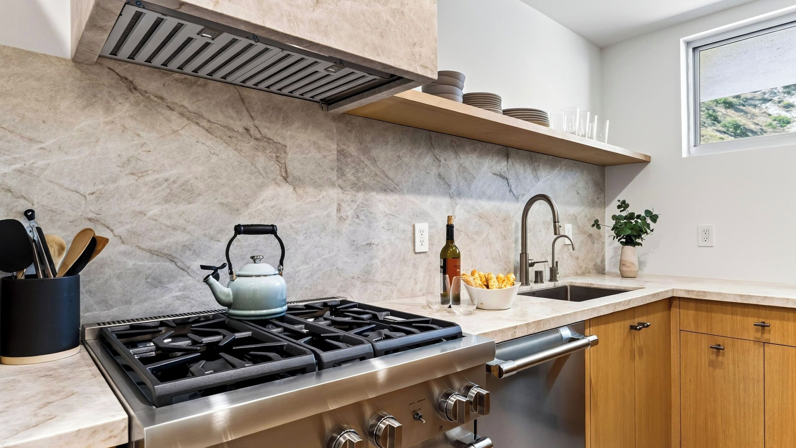 Una cucina moderna con piano cottura a gas, elettrodomestici in acciaio inossidabile, zoccolo di marmo e una finestra vista.