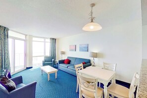 Een hotelkamer met een blauw tapijt, een bank, een eettafel met stoelen, een lamp en een raam met gordijnen.