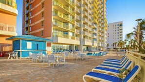 A hotel pool area with lounge chairs, a small blue building, and a large multi-story building in the background. - Bay Watch Resort 1512 (North Myrtle Beach)