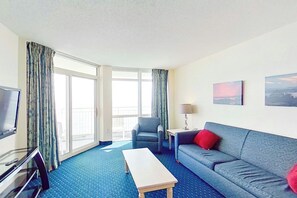 A hotel room with a sofa, a small table, a television, and a window with curtains. - Bay Watch Resort 1512 (North Myrtle Beach)