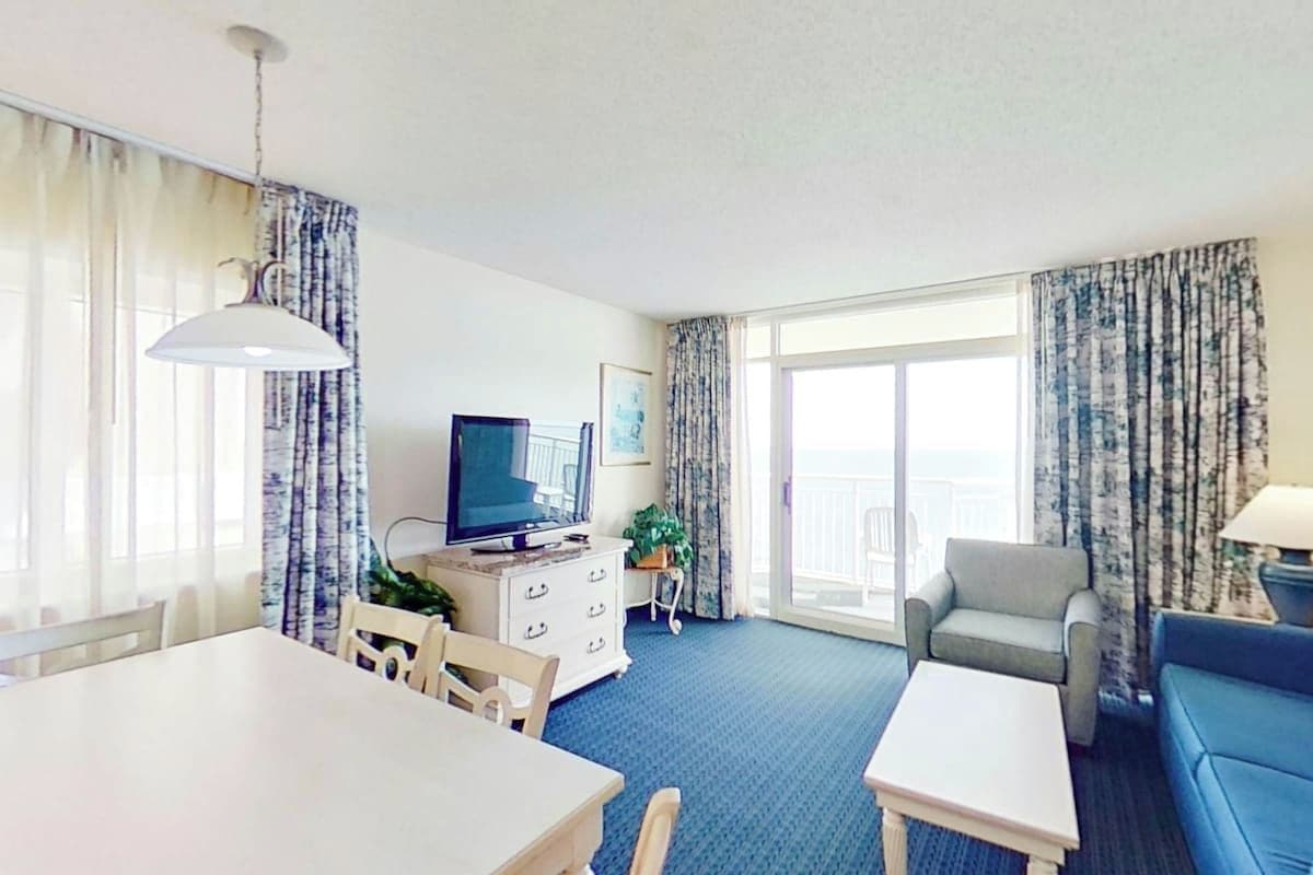 A hotel room with a dining table, a sofa, a television, and a balcony with curtains.