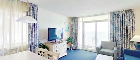A hotel room with a dining table, a sofa, a television, and a balcony with curtains.