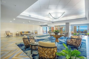 A spacious lobby with a high ceiling, a central seating area with wicker chairs and a glass-top table, and a view of the ocean through large windows. - Bay Watch Resort 1611 (North Myrtle Beach)