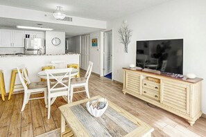 A modern kitchen with white cabinets, a dining area with a round table and chairs, and a TV on a wooden cabinet. - Bay Watch Resort 1612 (North Myrtle Beach)